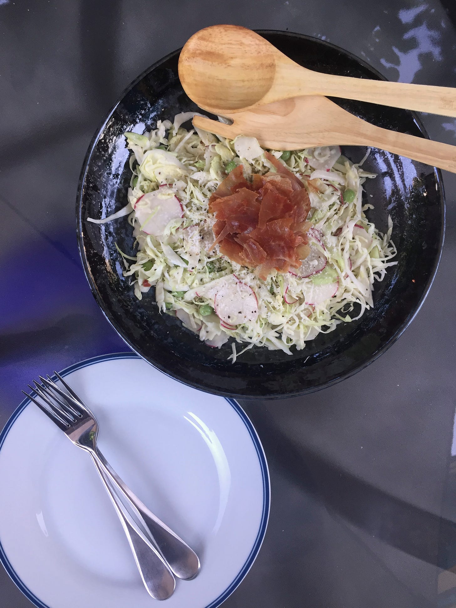 On an outdoor glass table, a shallow black dish of thinly sliced cabbages and radishes in a creamy dressing, with large green peas peppered throughout. A small pile of crumbled prosciutto sits at the centre, and salad utensils rest at the edge of the bowl. Two salad plates with forks on top are in the bottom left corner of the photo.