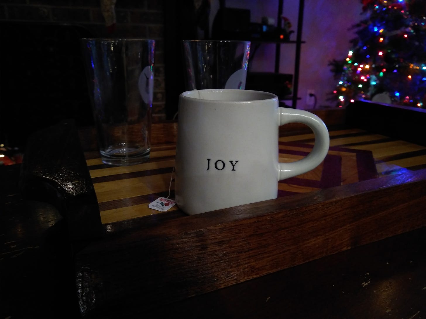 A mug that says JOY on a wooden tray, Christmas tree in corner background