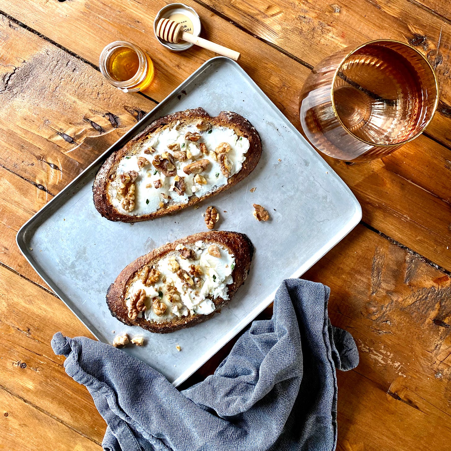Aluminium baking tray topped with two slices of toast covered in goats cheese, walnuts and honey. A glass of water in top right and a small jar of honey to the left. 