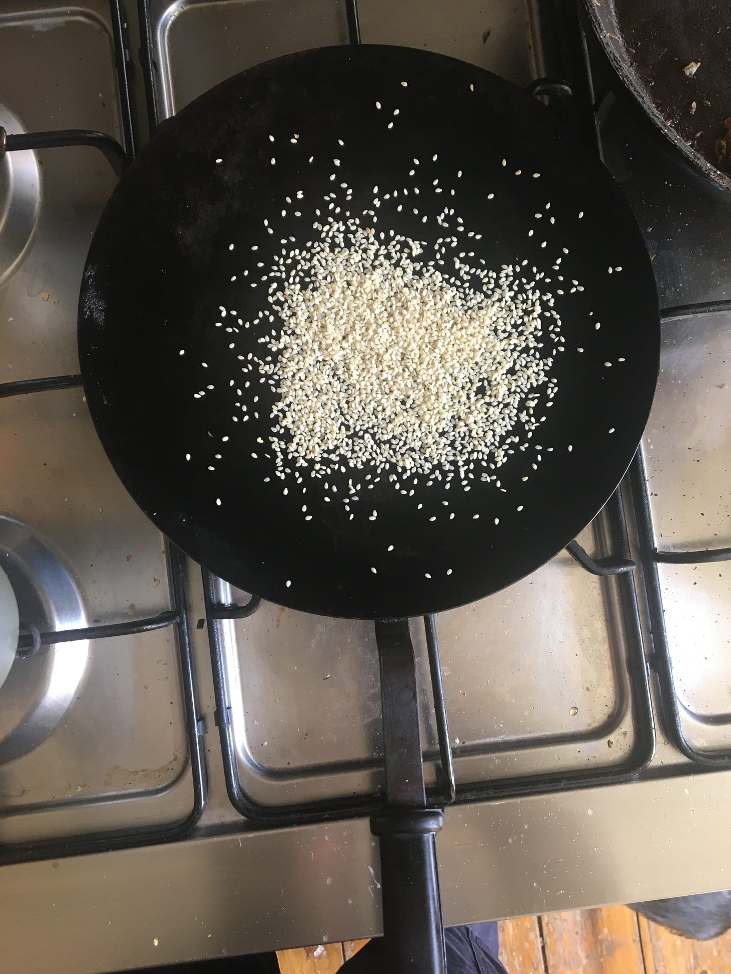 A flat round iron pan photographed from above, on a semi-clean metal gas hob, on which are toasting a pile of pale sesame seeds. The pan has a long dark handle that is in the foreground. 