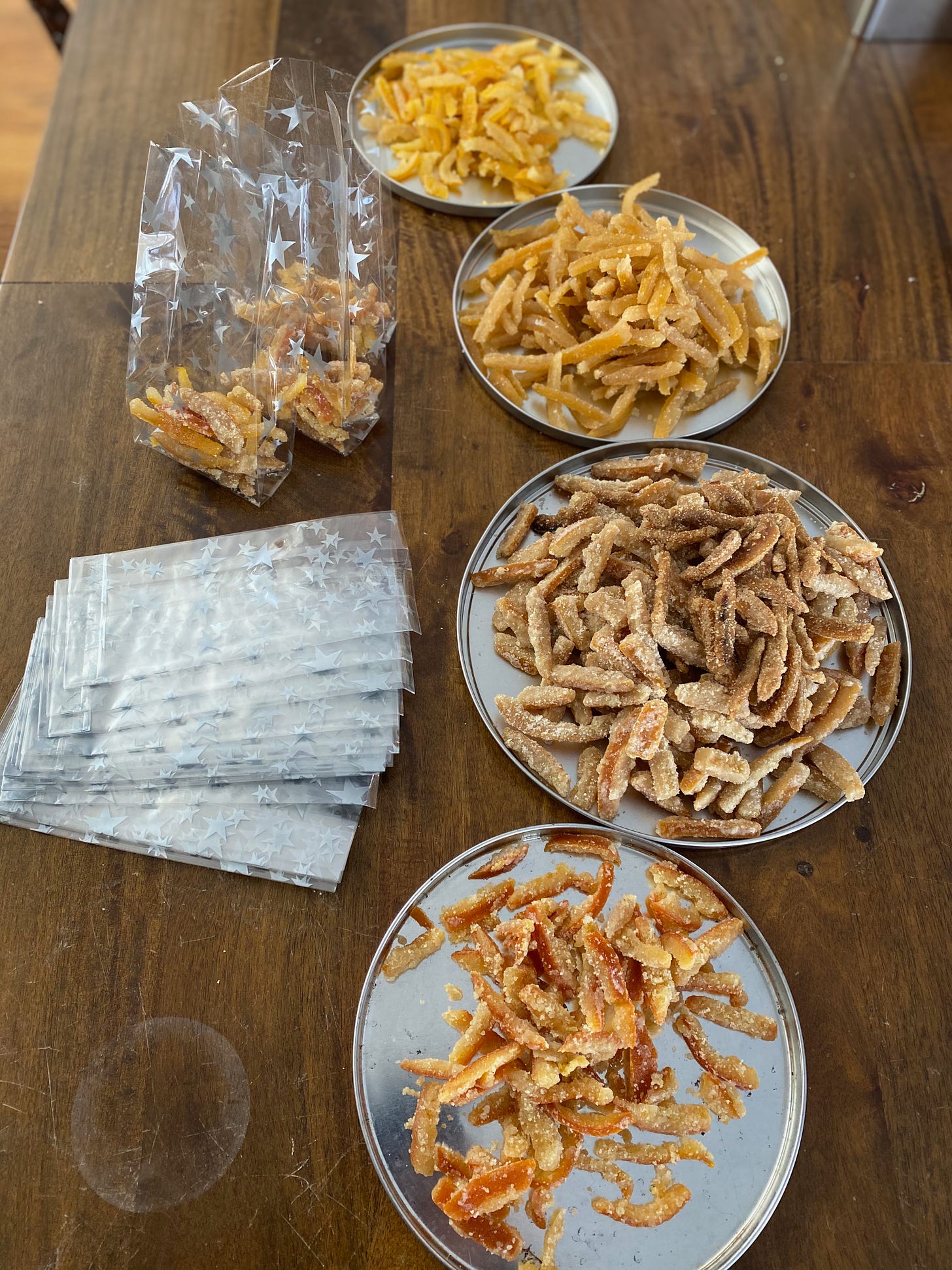 Four plates of candied peel—Meyer lemon, lemon, orange, and blood orange—sit on a wooden table. There are also two small clear gift bags partially filled with peel, and a big stack of unfilled gift bags.