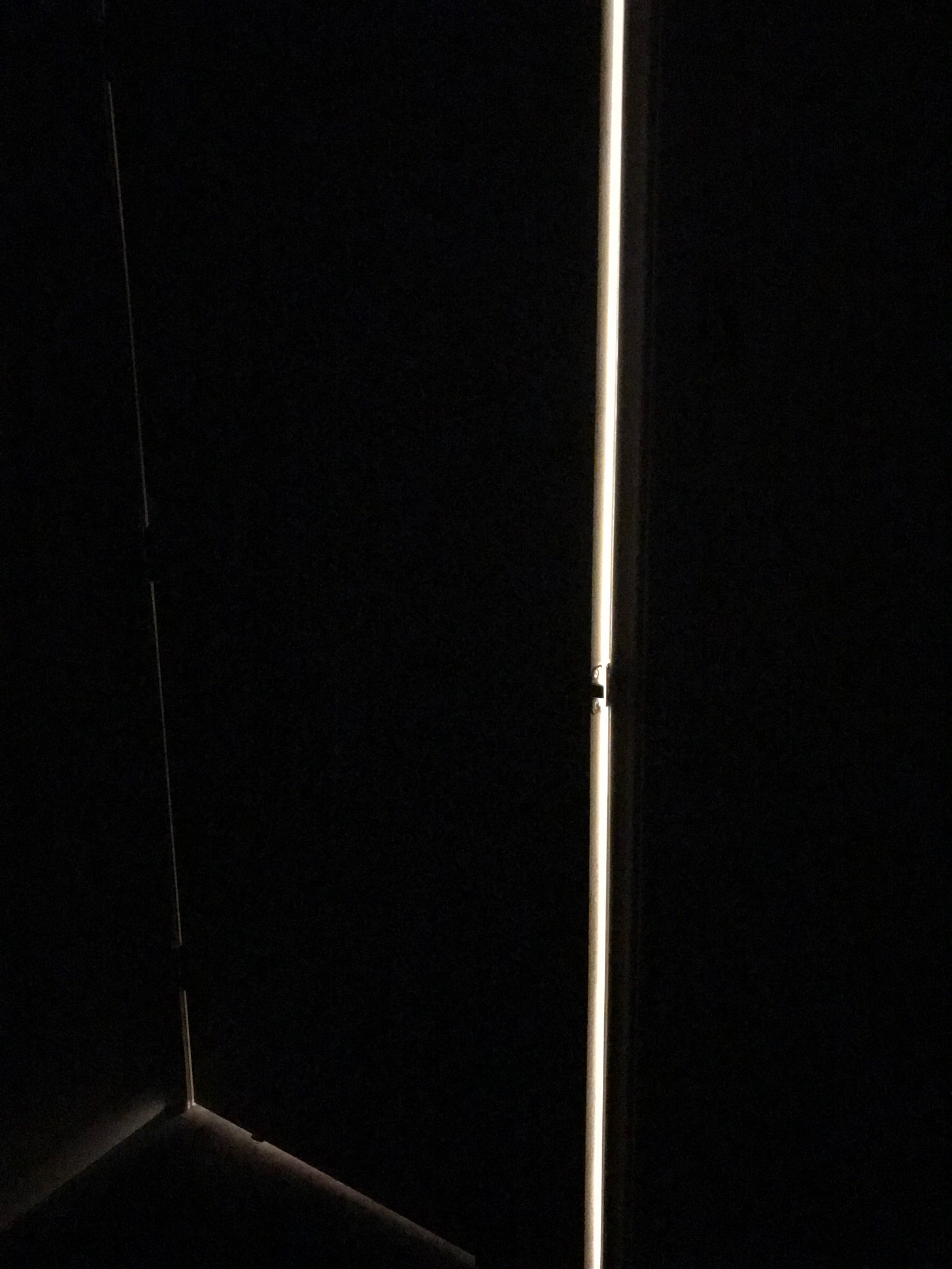A dark room holds the faint outline of a cracked door. A think vertical line of light shine through.