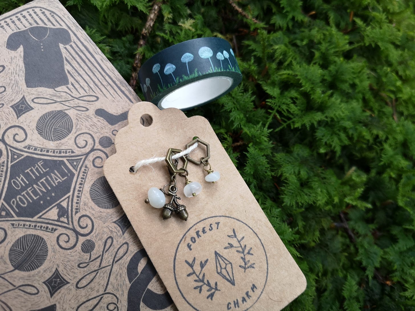 Image description: A bundle of goodies lying on some squishy green moss: a set of stitch markers and progress keepers featuring moonstone beads and an acorn charm, lying on top of crafting notebook and next to a roll of dark blue washi tape with white mushrooms.
