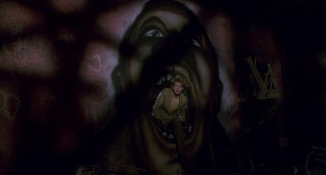 Virginia Madsen emerges from a hole in the wall, which is also  a graffitti mouth of a black man