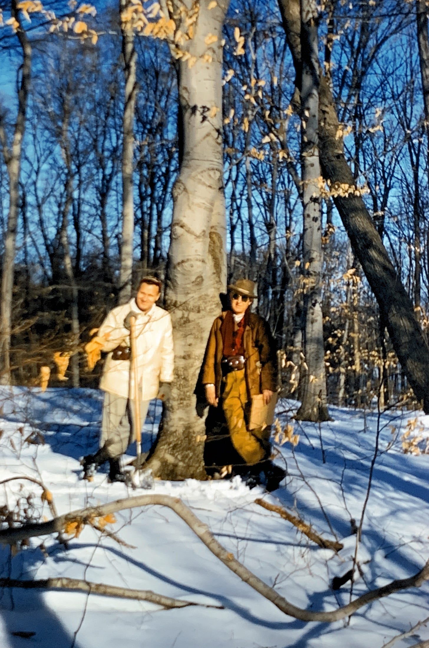Two men standing next to a tree with smooth bark. There is snow on the ground and they are wearing winter coats and gloves.