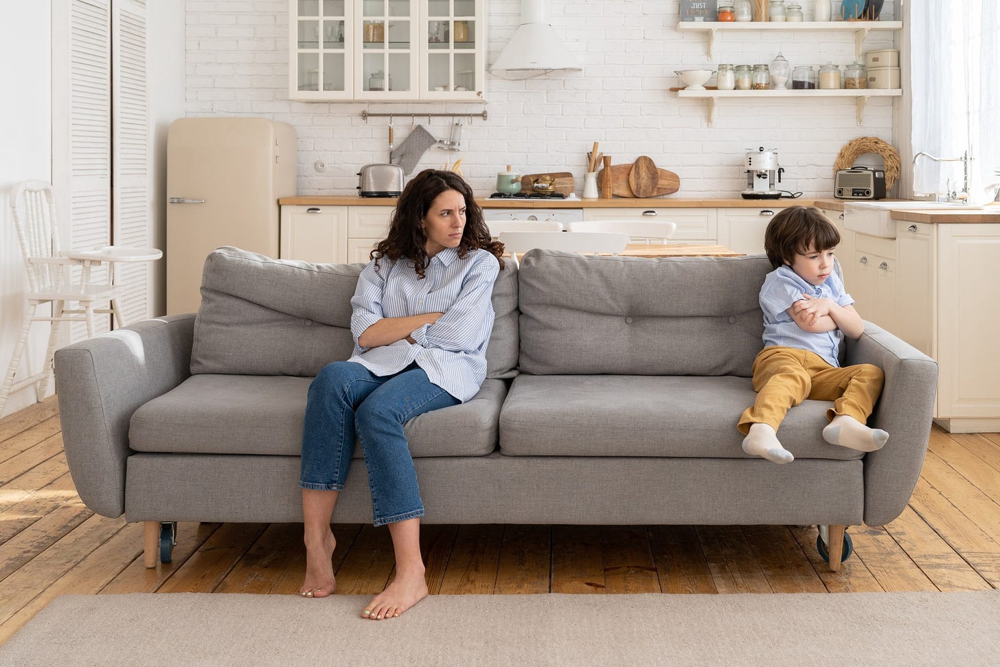 A mother glares at her child as they sit with their arms crossed on opposite ends of the couch.