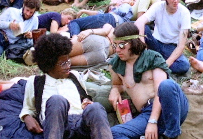 Black hippie with an Afro talking to a white long-haired hippie at a festival in the ‘60s