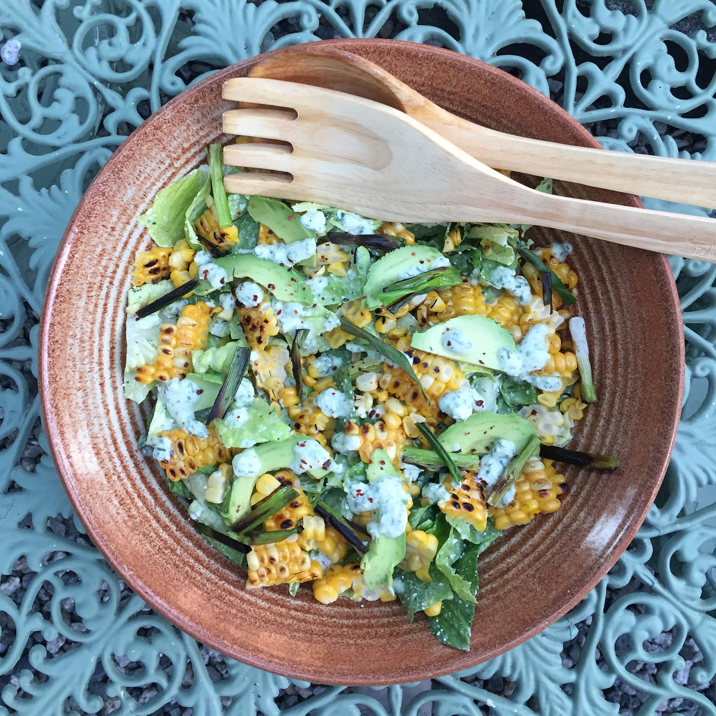 from above, a rustic ceramic bowl filled with salad greens, slices of avocado, bits of grilled green onion, and large pieces of charred corn sliced off the cob. A creamy dressing is drizzled over the top. A wooden serving fork and spoon sit near the top of the bowl.