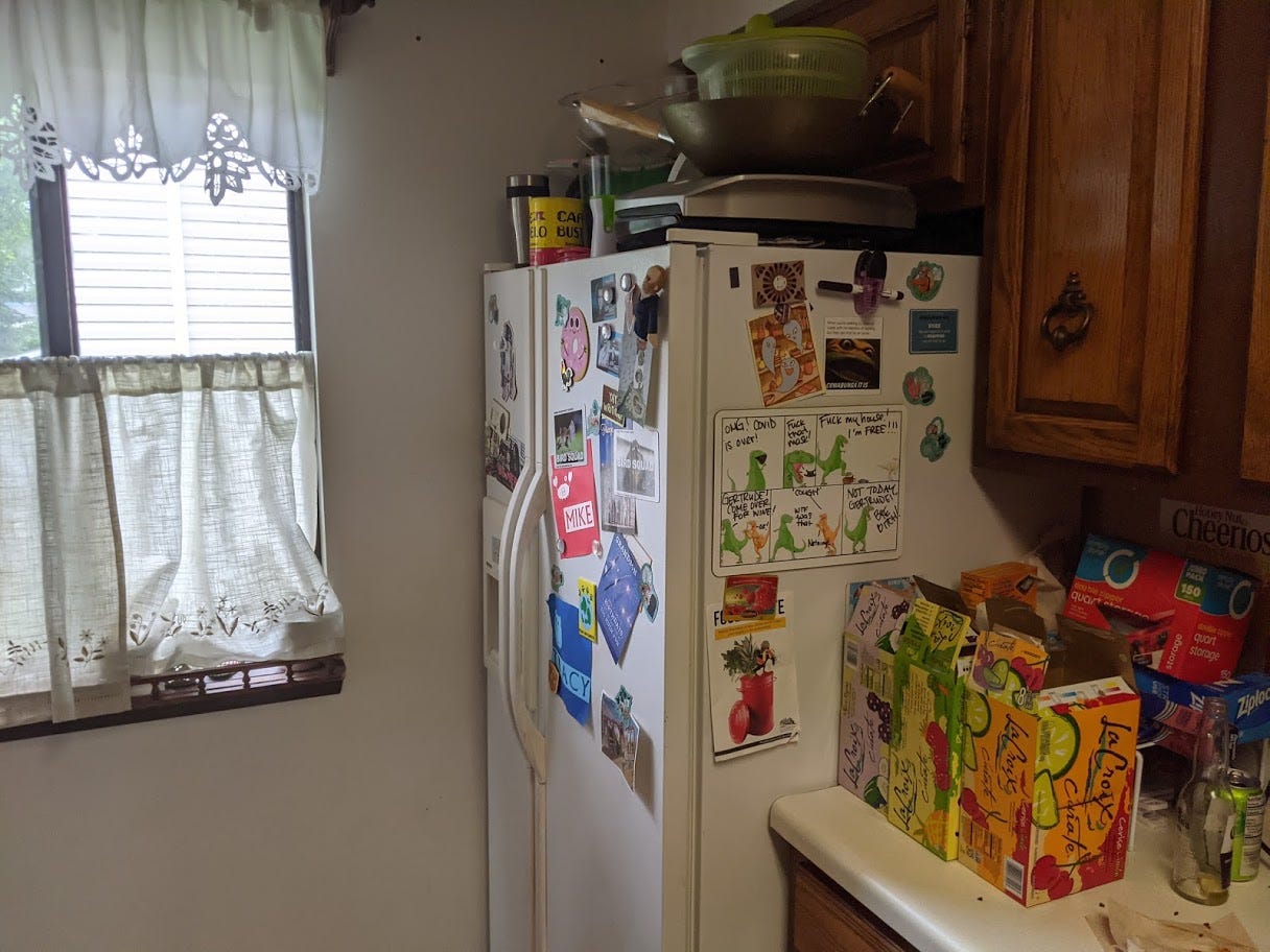 A fridge with various magnets stuck to the front, kitchen items stacked on top, and boxes of La Croix on the counter next to it, as well as an empty bottle and can.
