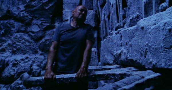 John Locke (Terry O'Quinn) grasps the spoke of an enormous wooden wheel embedded in the rock wall of a cave, and struggles to move it.