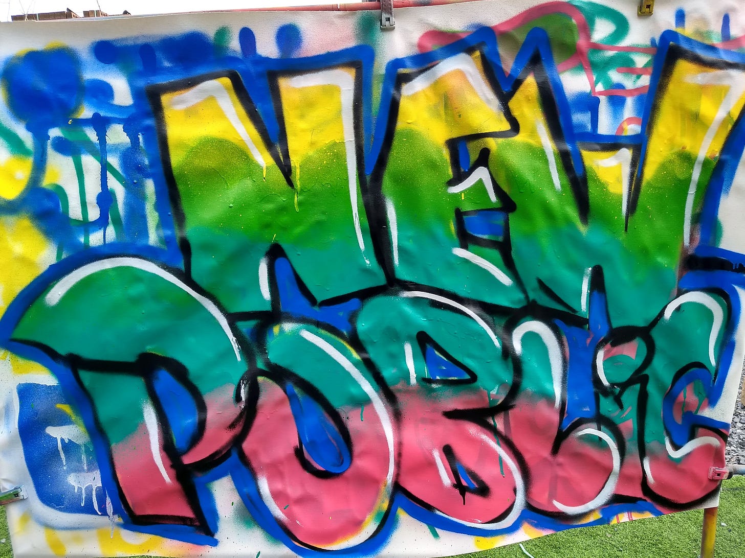 A physical canvas spray painted with big bubble letters reading “New_ Public” in yellow, two greens and pink, with blue, black and white as accents.