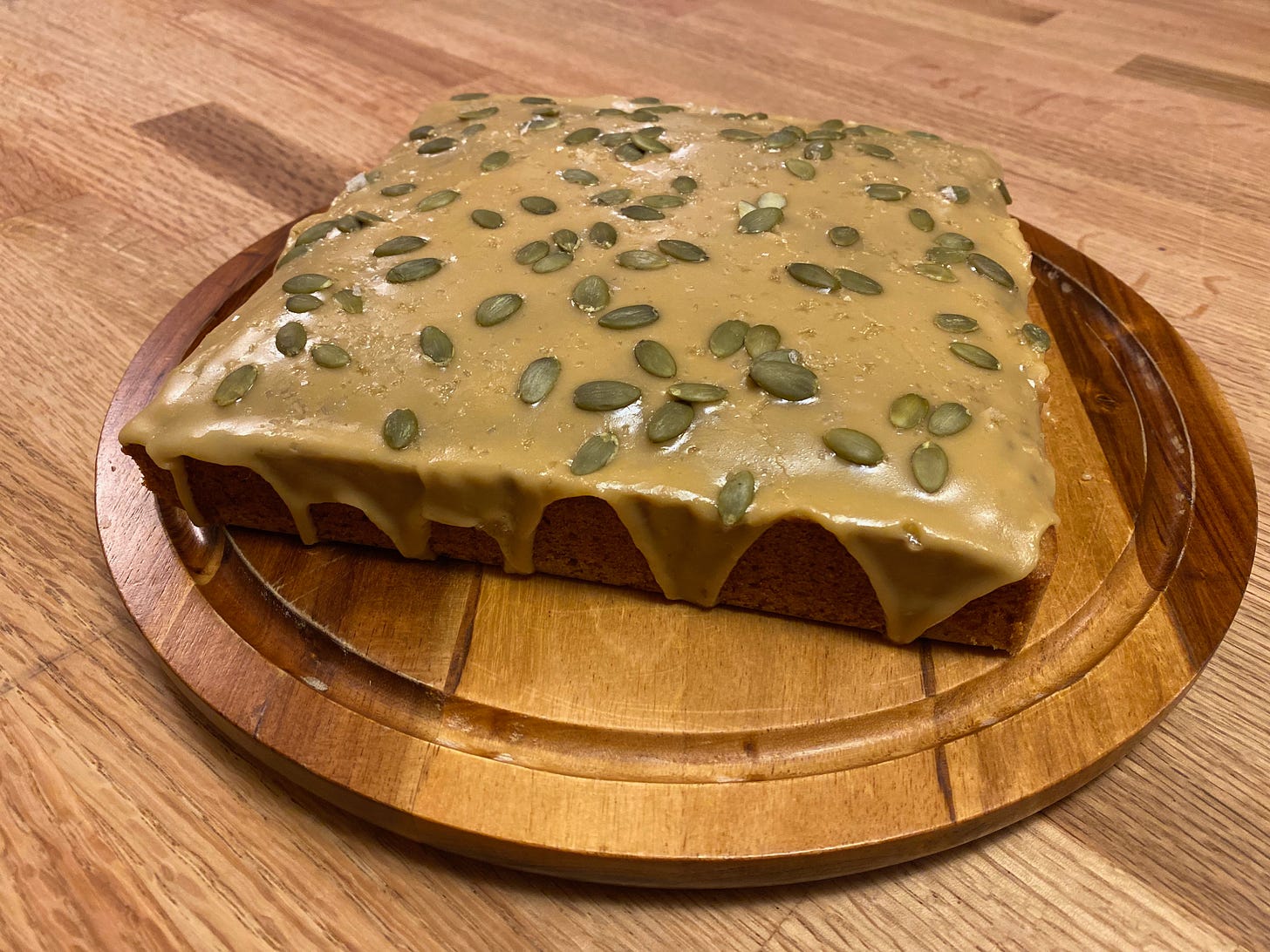 A square cake glazed with a shiny caramel-colored frosting and scattered with pepitas and flaky salt.