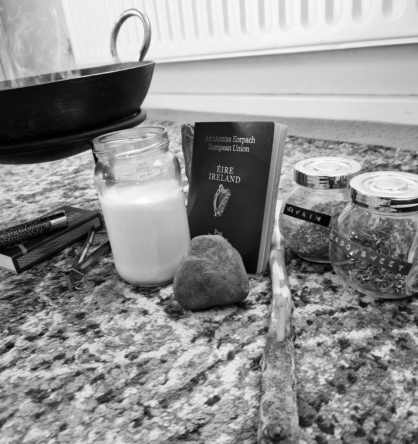 A black and white photo of items on a rug on the floor. The items are a cast iron pot with smoke coming out of it, a lit candle, a lighter, an Irish passport, a large stone, a homemade wand and two pots with dried Vervain and Meadowsweet respectively.