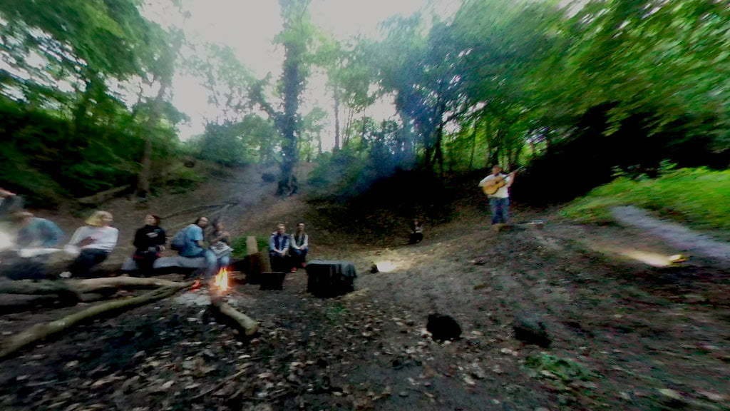 People sat around a fire in an abandoned chalk pit. A man stands and plays guitar.