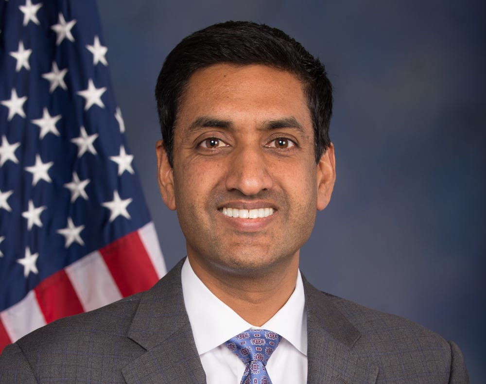 A headshot photo of a middle-aged man of Indian descent in a suit in front of a dark blue background and American flag.
