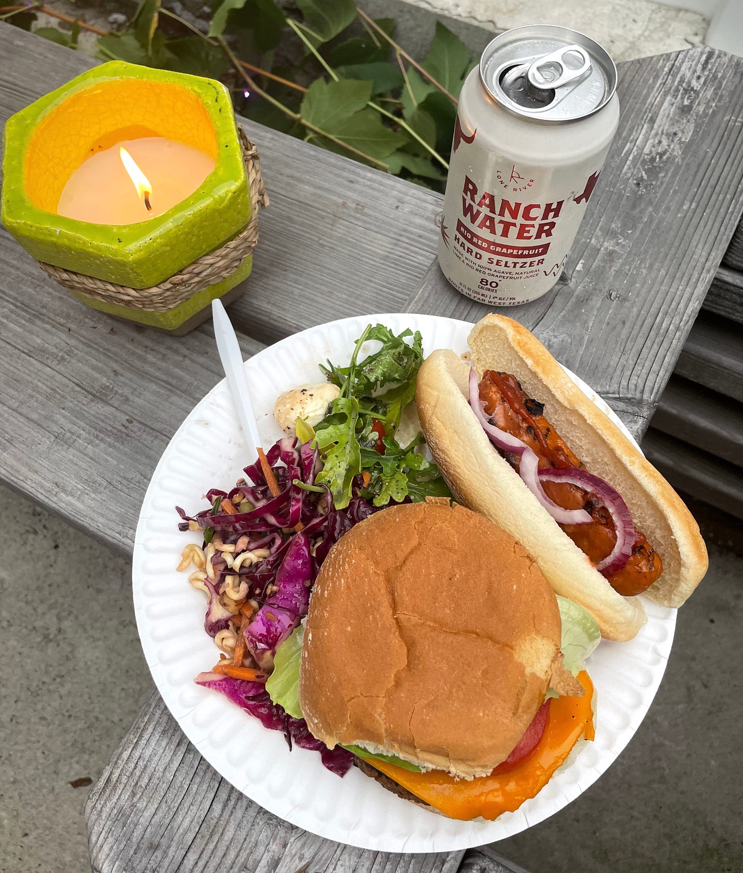 plate with veggie burger, slaw, salad, veggie dog next to candle and hard seltzer