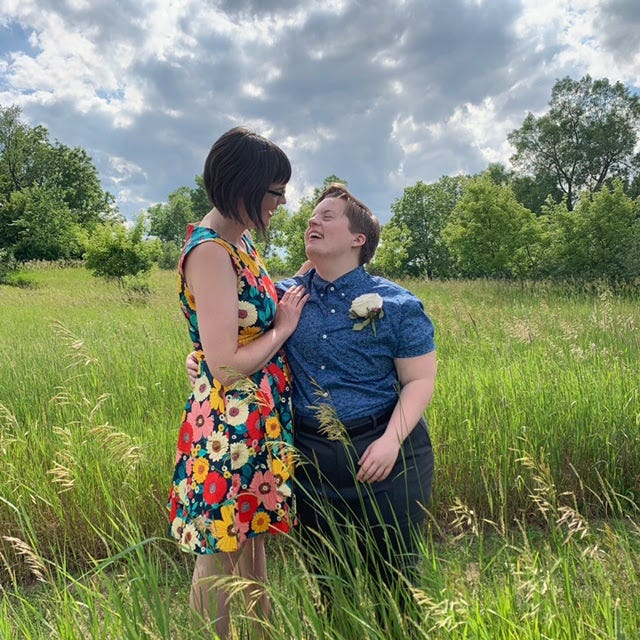 Heather and Victoria stand in a field on a sunny day with their arms around each other and smiling at each other. Victoria has short brown hair and is wearing a bright floral sundress. Heather has short light hair and is wearing a blue button-down, jeans, and a boutonniere.