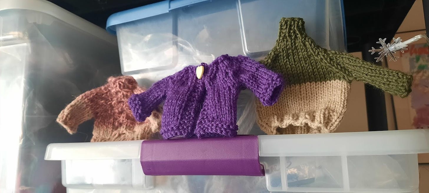 Three sweaters in pink & brown, purple with a yellow button, and light and dark green as holiday decoration on a box.
