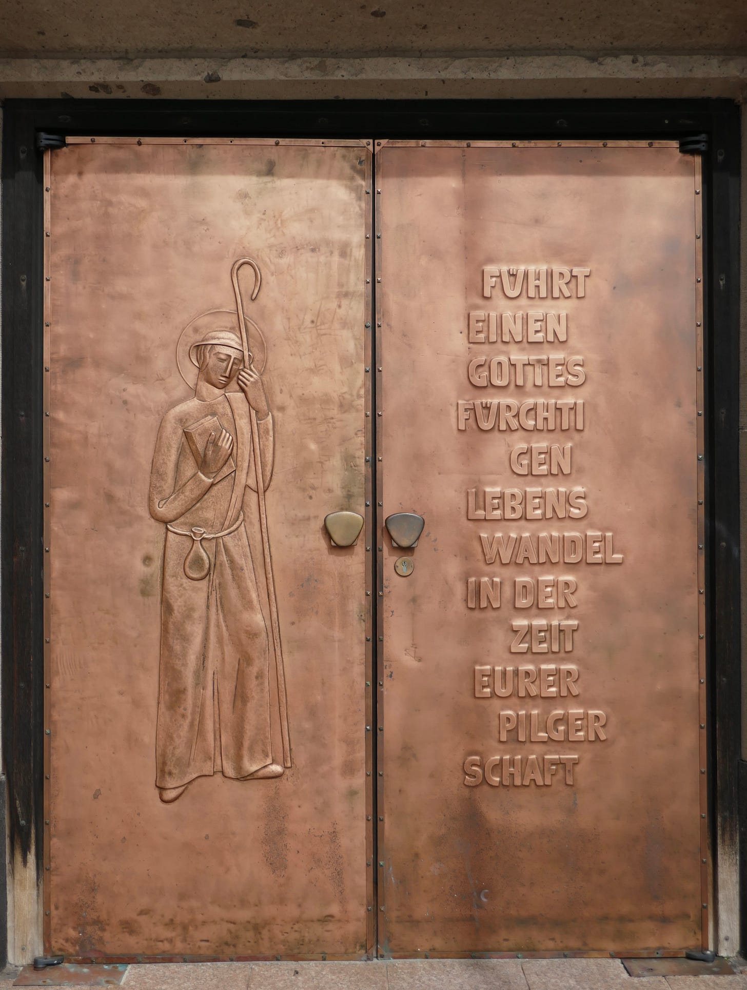A double copper door in Cochem, Germany. One side has a relief of a pilgrim and the other side a message in German, “lead a god-fearing walk of life during the time of your pilgrimage.”