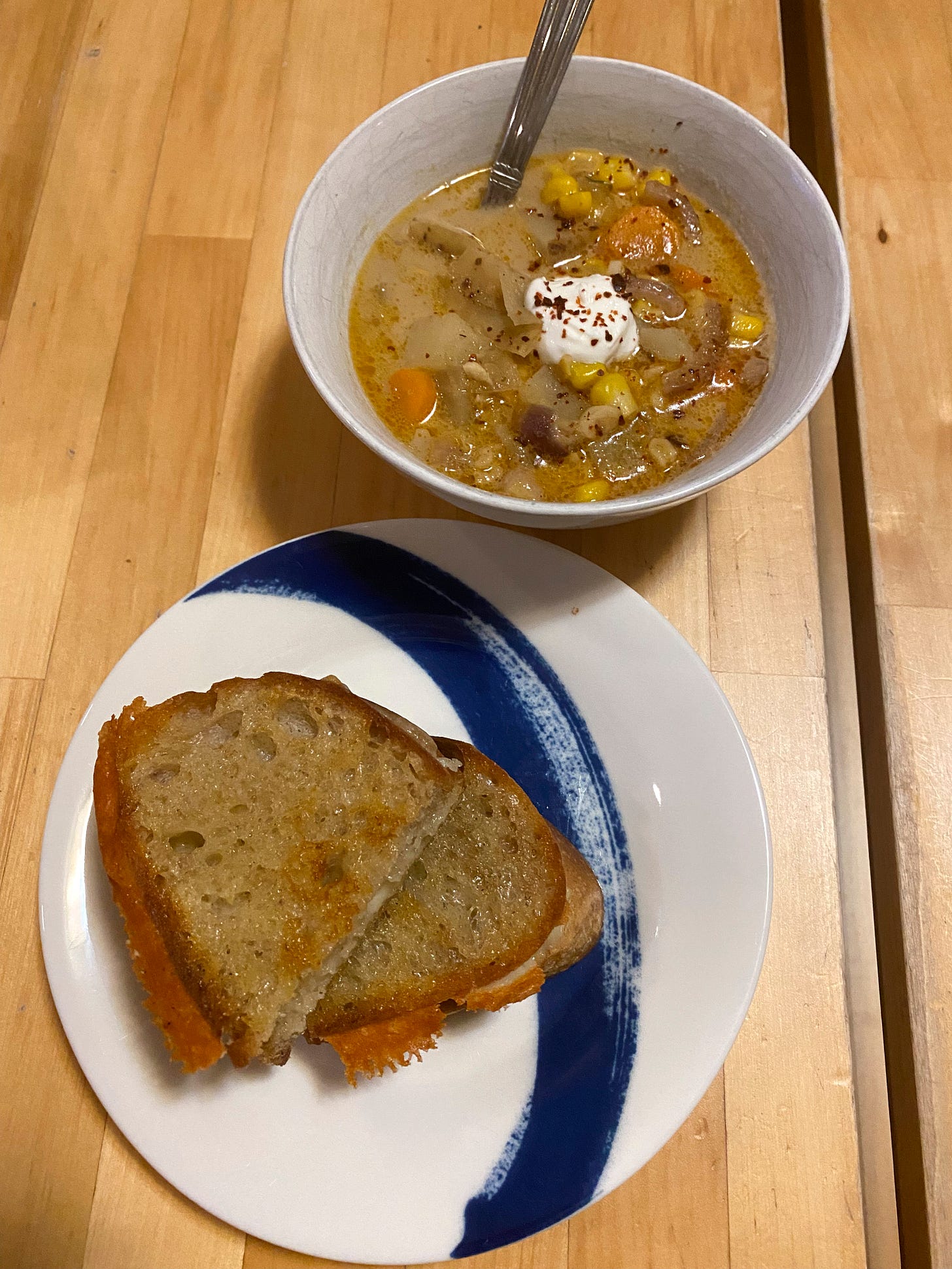 A small blue and white plate with two halves of a grilled cheese sandwich is in the foreground. Cheese has formed a crispy lattice at one edge of the bread. Behind the plate is a small white bowl filled with corn chowder, with chili flakes and a dollop of yogurt on top.