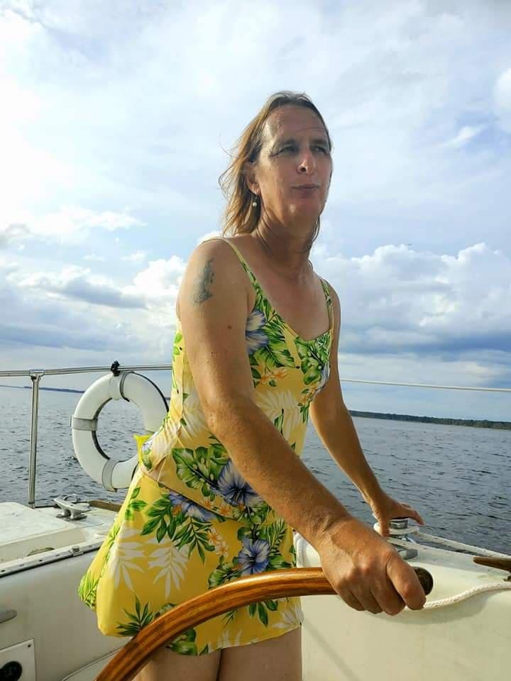 A woman is standing at the helm of a boat, tiller in hand. Her long hair flows behind her, she is in a yellow floral top and skirt. 