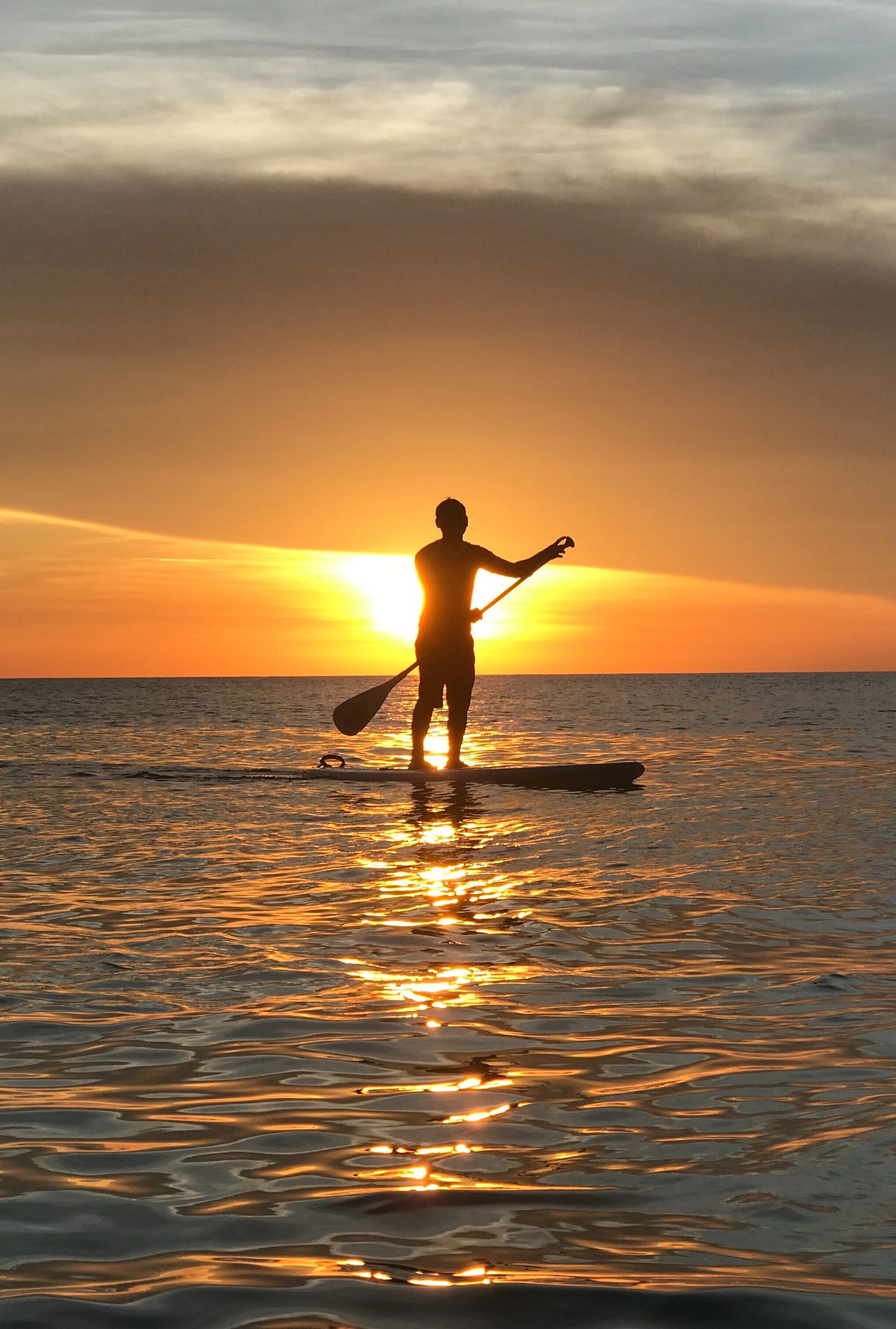 individual on a sea board silhouetted in front of the glowing sun floating on waters reflecting the sunlight.