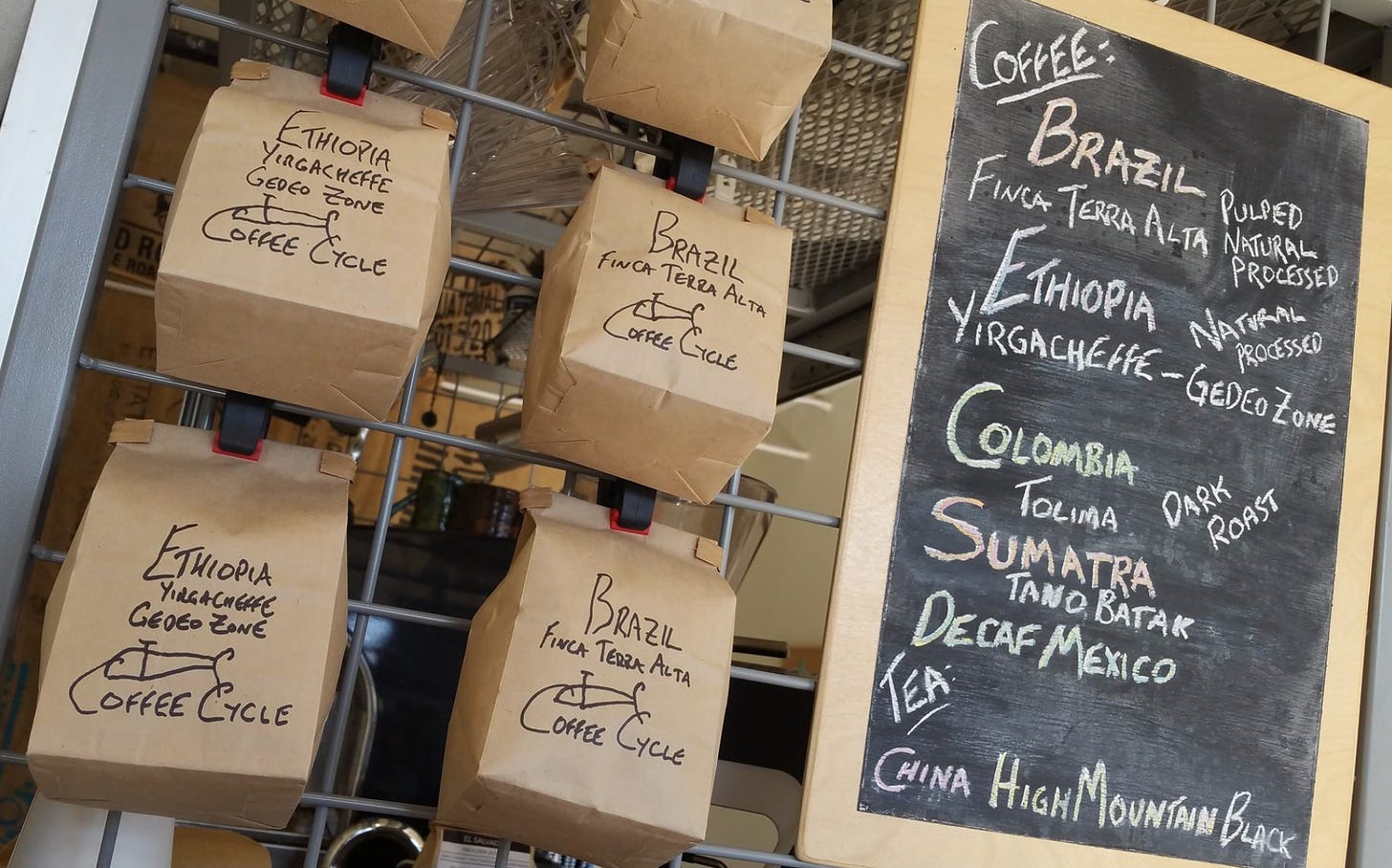 Handwritten Coffee Cycle brand coffee bags hanging on a cage next to a chalkboard with the different types of coffee available listed vertically.