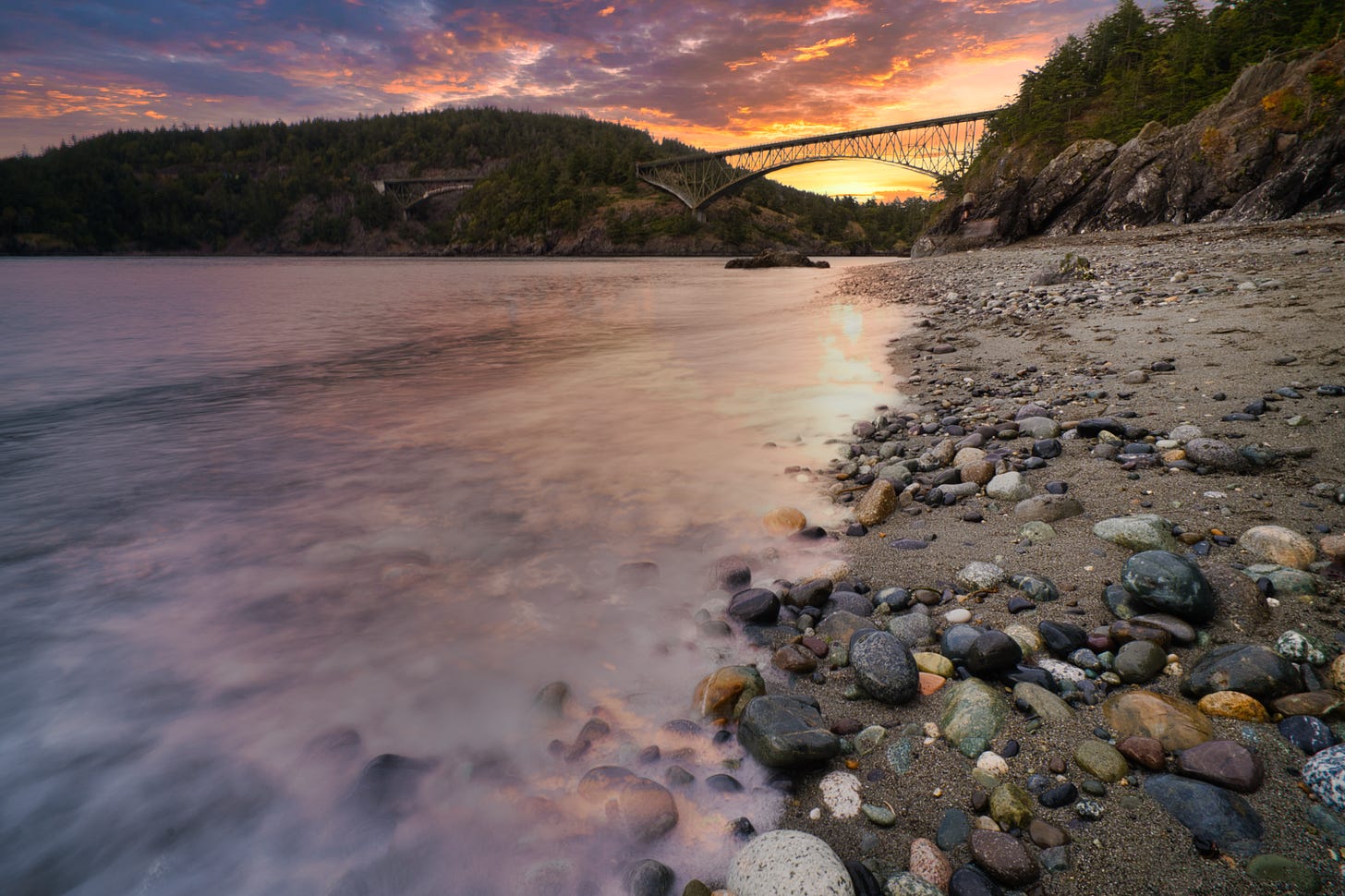 A beach at sunset, with sand and rocks at right and water at left, looking toward the Deception Pass Bridge in Washington state.