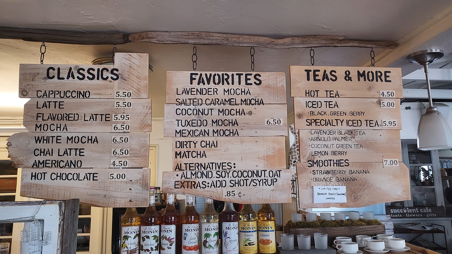 Photo of a hanging scrap wood menu sign at a cafe. Raw wood with black, stencil lettering. A line-up of coffee syrup bottles is under the menu at the bottom of the frame.