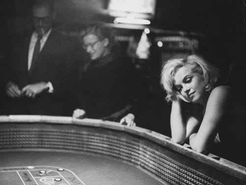 foto em preto e branco, da atriz marilin monroe ao canto direito, olhando para uma mesa de jogos, com a cabeça apoiada na mão direita.