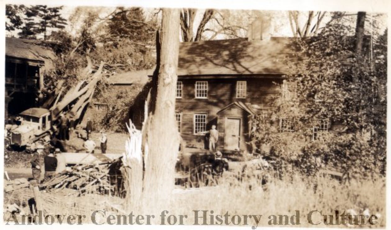 sepia-toned photo of brown-sided house with tree limb resting on shed roof. Men stand by with truck. 