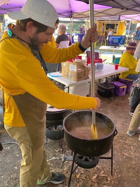 Zach, a thin man in a white baseball cap, long-sleeve gold shirt, tan pants and apron, stirs a large pot of food with a metal paddle.