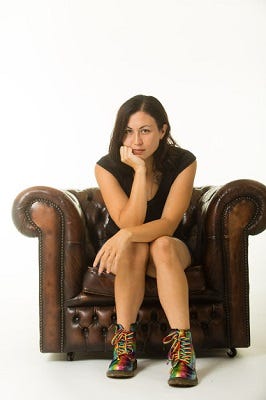 Picture of Sunyi Dean, a brown-haired woman sitting in a thoughtful pose in a large brown armchair, against a white background. She is wearing black clothing and rainbow colored boots.