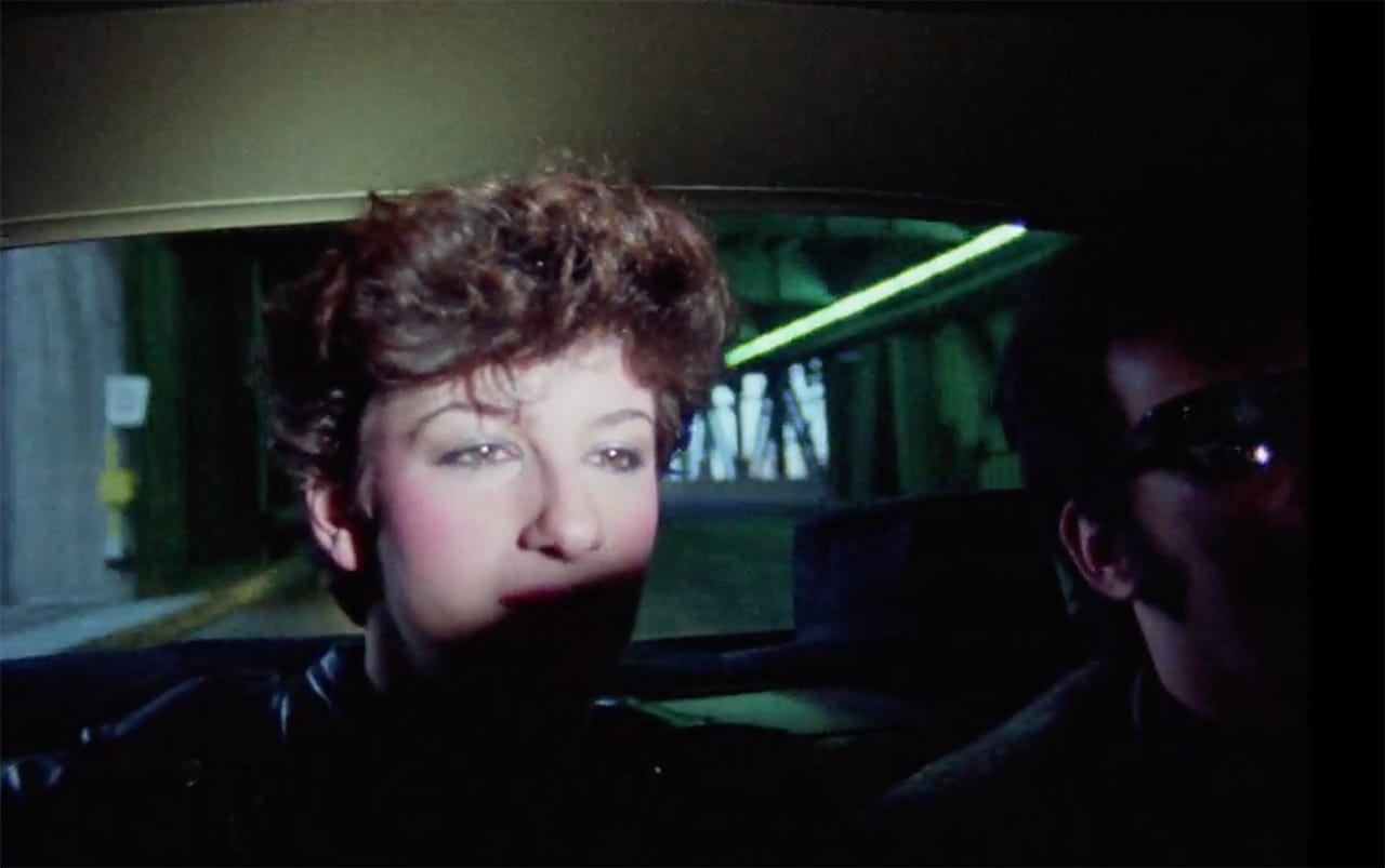 Sharon Mitchell riding in the back seat of a car with the camera light spilling over her forehead in Kamikaze Hearts (1986)