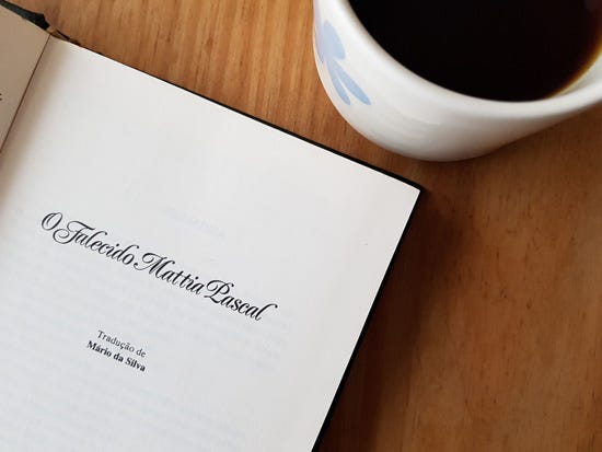 Foto colorida do livro O Falecido Mattia Pascal aberto numa página que mostra seu título. O livro está numa mesa de madeira ao lado de uma xícara de café.