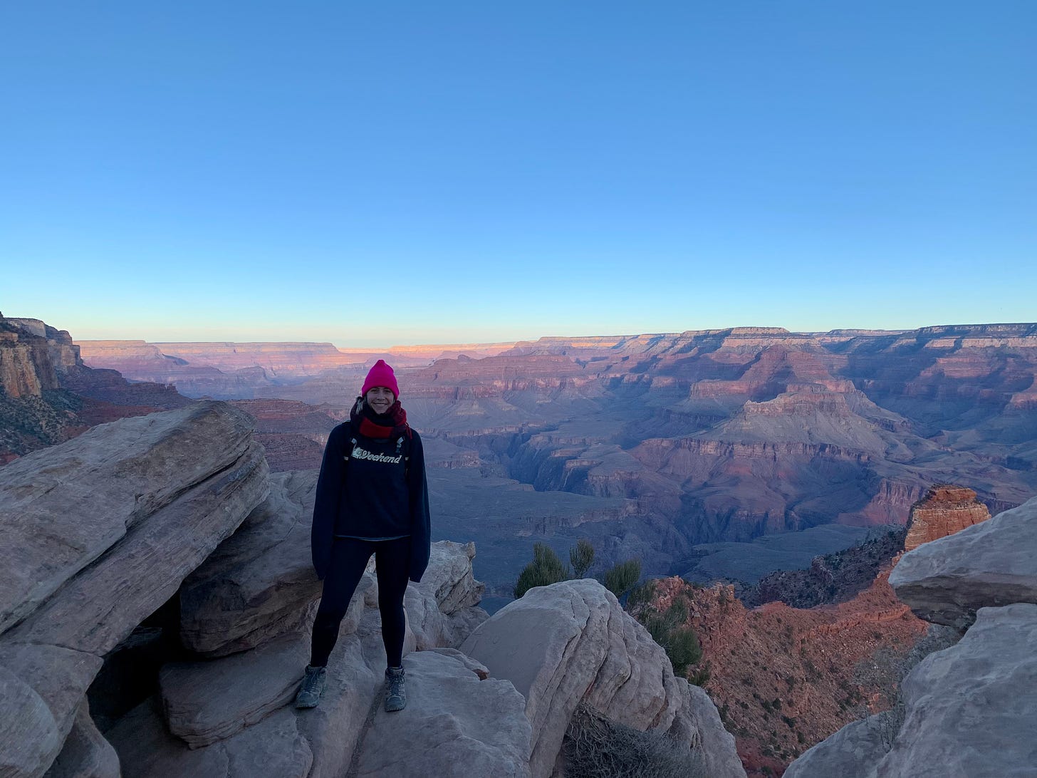 A woman stands above the Grand Canyon at dawn, a rainbow of colors appearing on the ragged landscape far below her