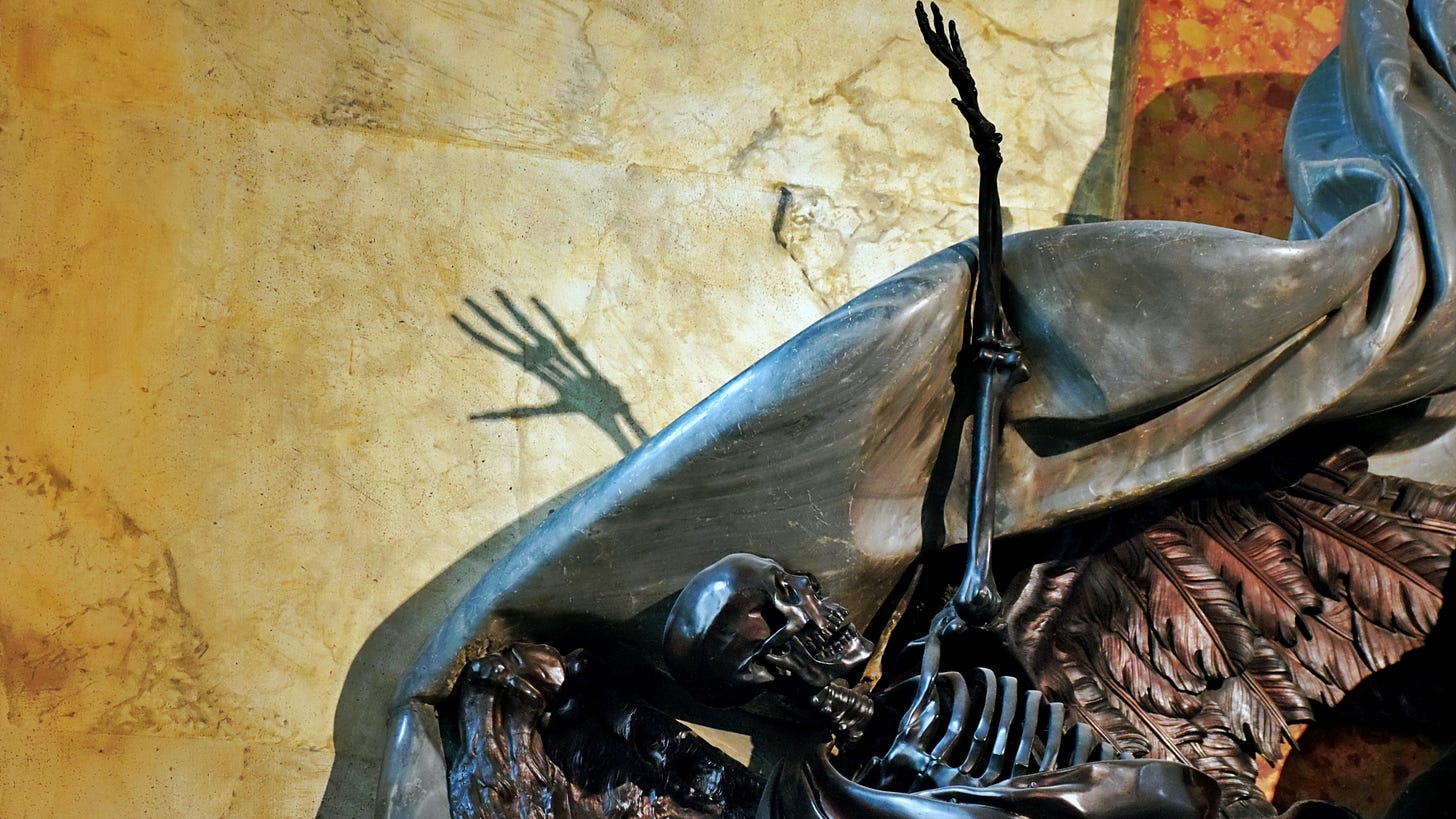 Sculpture of black skeleton with left arm reaching upwards, shadow of left hand on marble wall behind