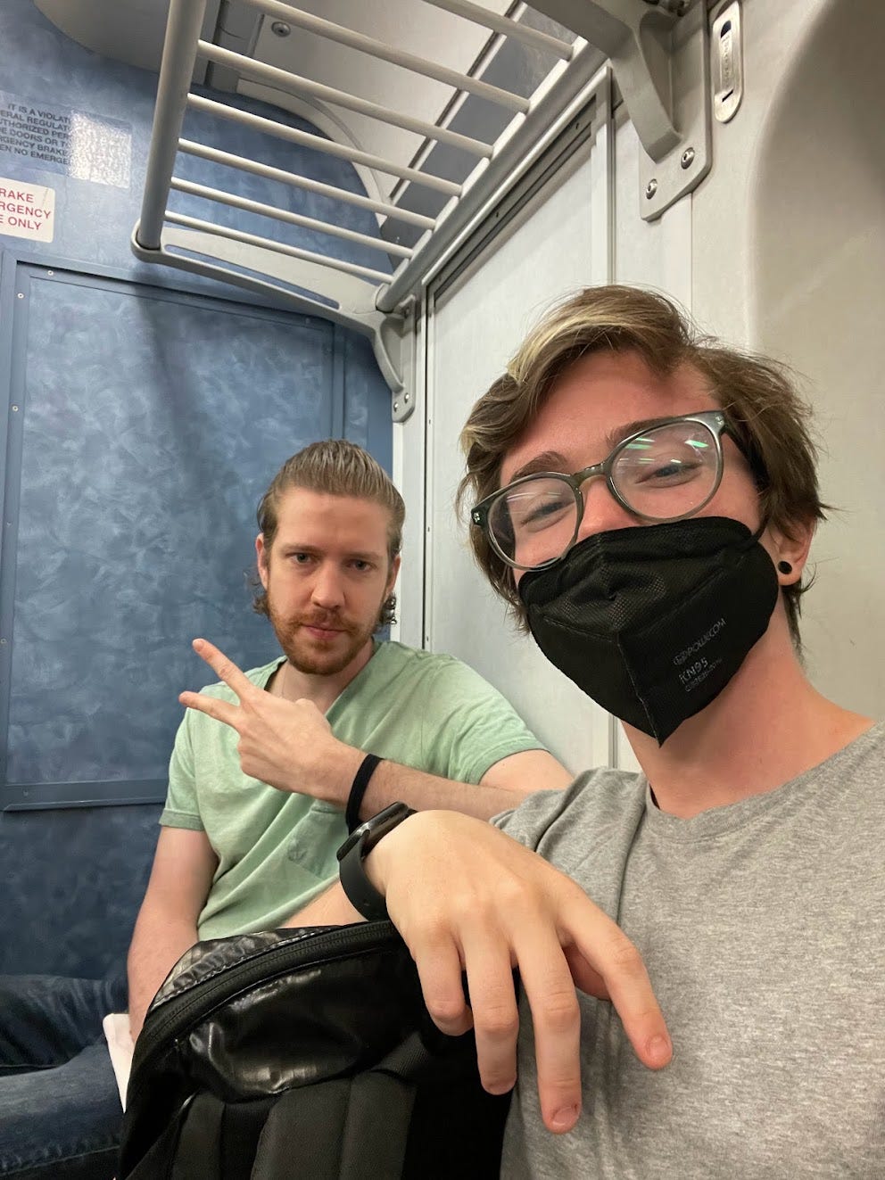 A blond white man makes a peace sign next to a brown-haired person wearing a mask on a train