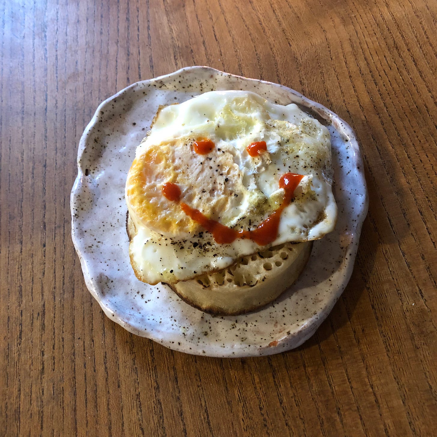 A fried egg atop a single crumpet, on a small place. A red smiley face is squirted over it in sriracha. 