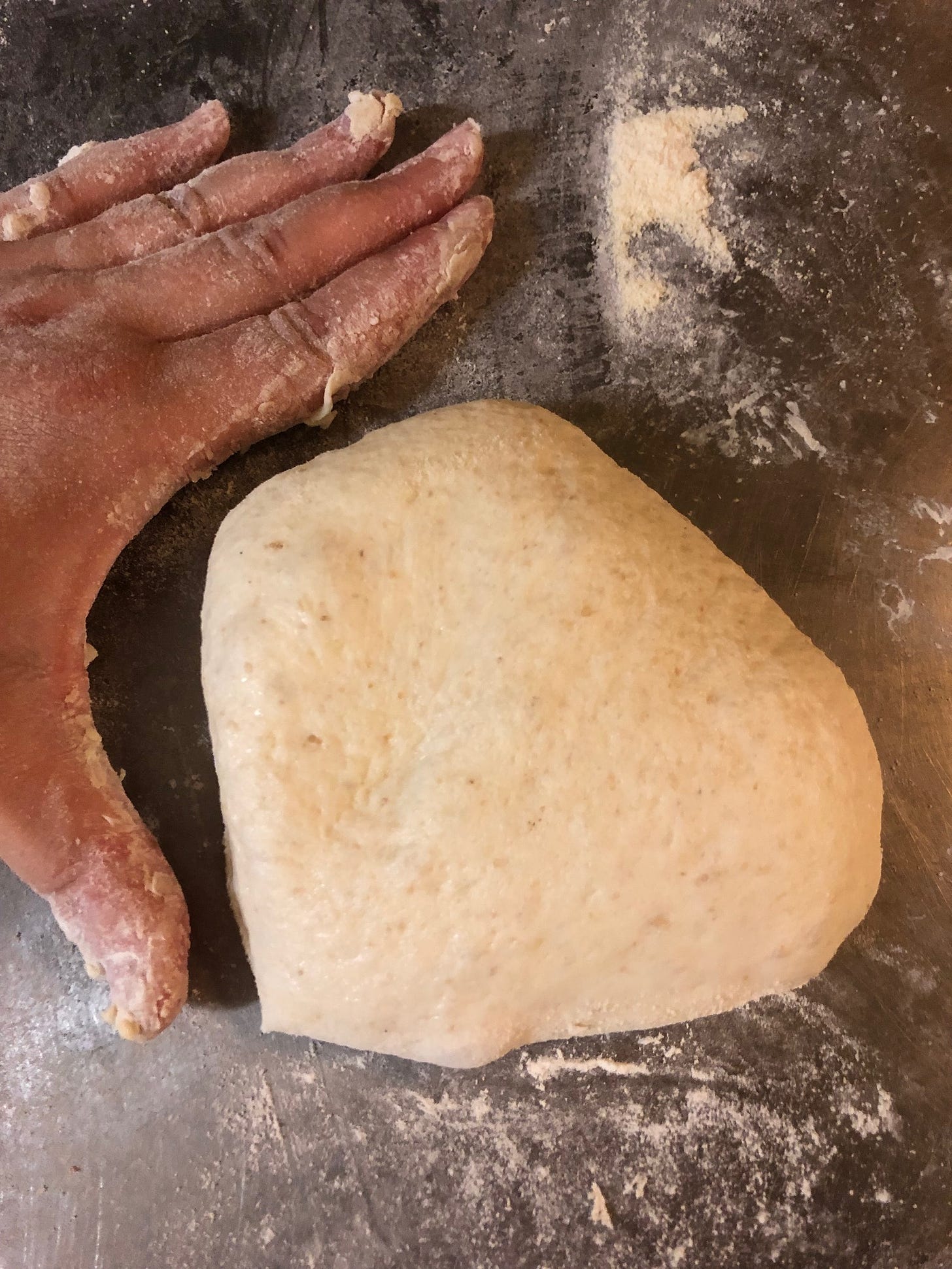 Left hand, palm down, on a metal table, next to a piece of raw unshaped dough.