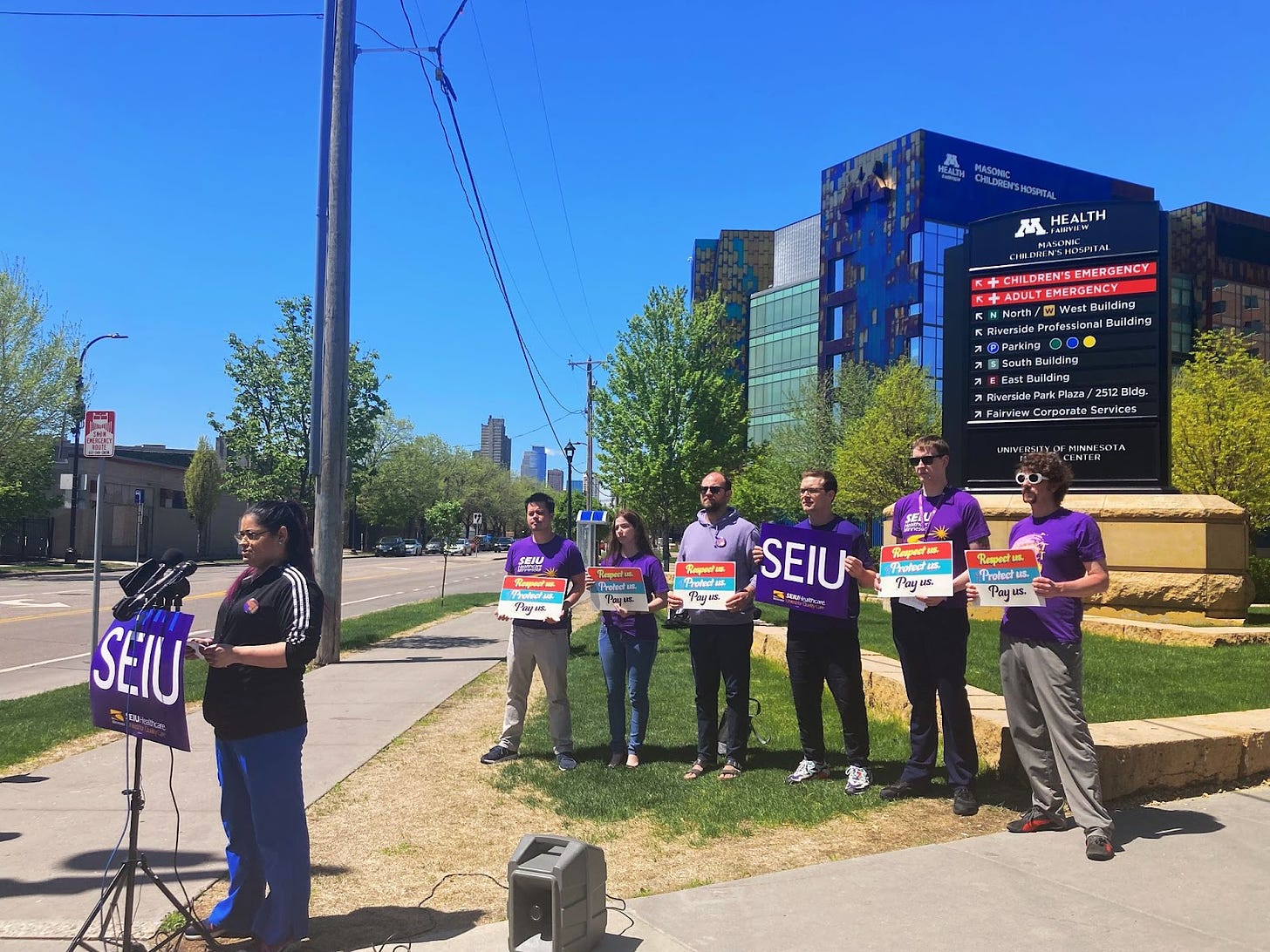 a small group of people wearing purple shirts and holding SEIU signs and signs saying "respect us protect us pay us" stand outside of a blue hospital building on the sidewalk