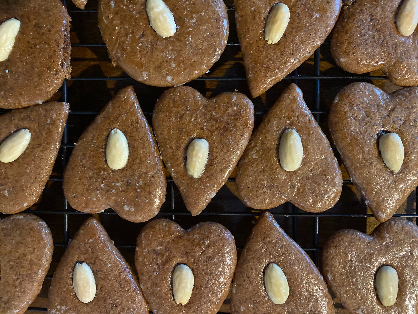 A closeup of many heart-shaped gingerbread cookies on a wire cooling rack. The cookies are covered with a shiny glaze and each one has a blanched almond pressed into its center.