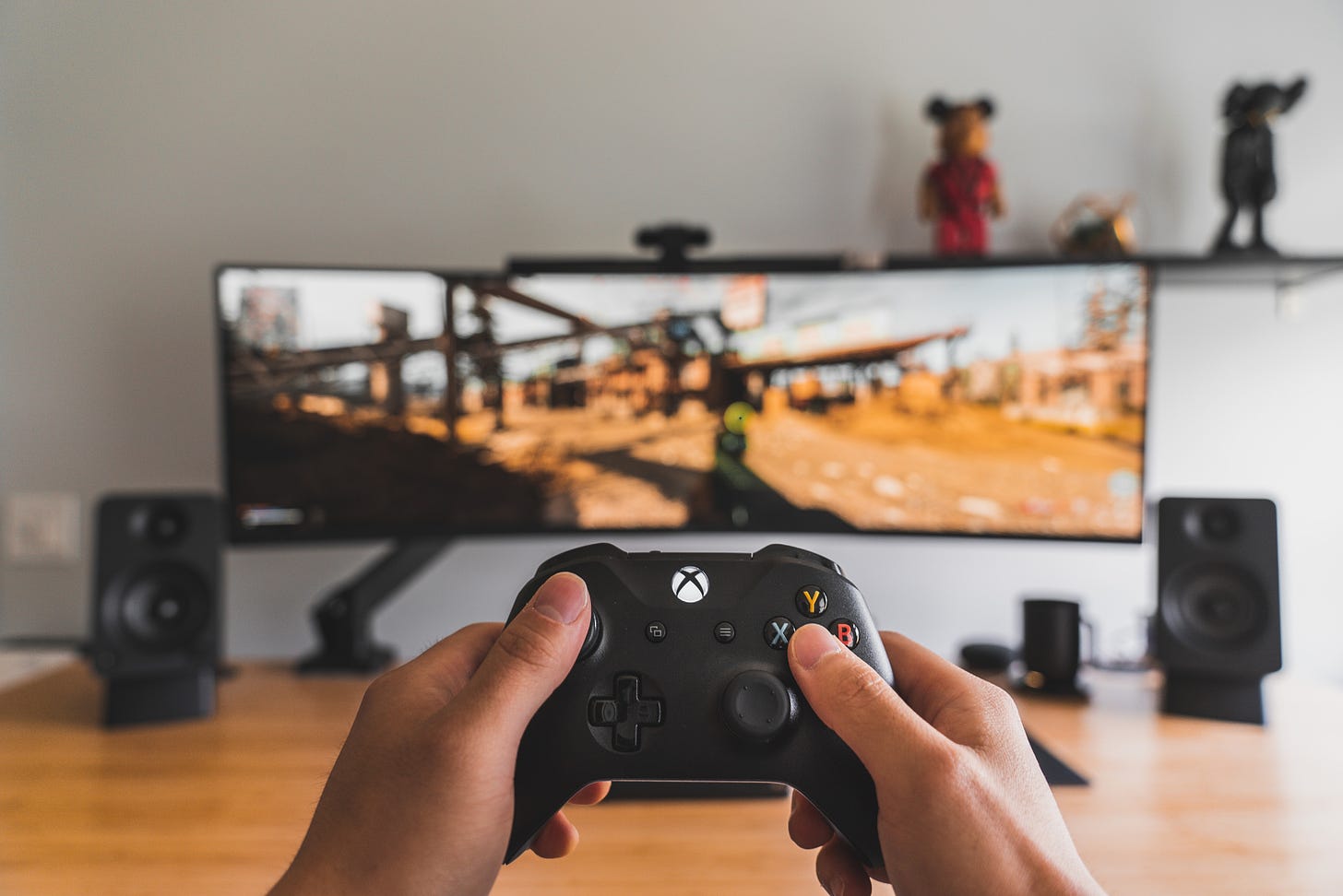 Hands holding a gaming controller towards a desk, monitor, speakers and a shelf