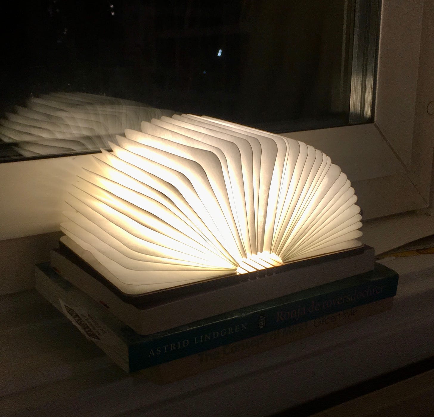 Auf einer Fensterbank liegen zwei Bücher, darauf eine kleine Lampe, die aussieht wie ein aufgeschlagenes Buch. Das ist die einzige Lichtquelle, sonst ist es dunkel.
