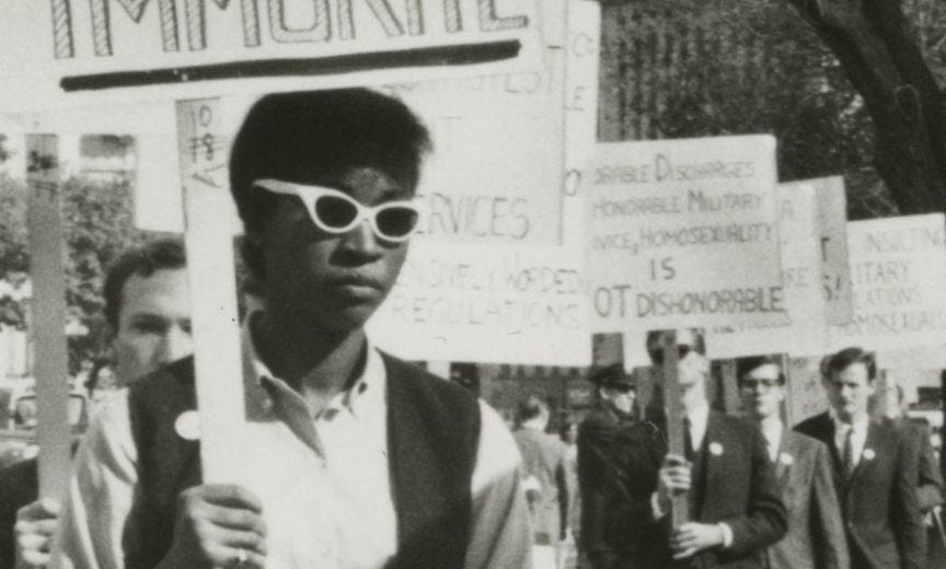 Ernestine Eckstein, a Black woman with white sunglasses, a button, and a sign, pickets at the Annual Reminder Day, leading a line of white men in suits.