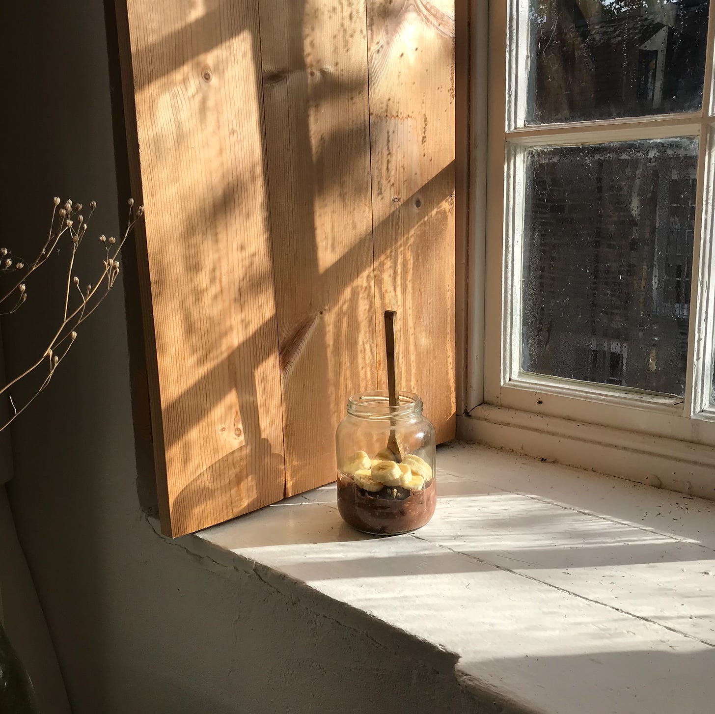 A window ledge in harsh bright morning light, with honey warm wood on the side panels. On the ledge is a clear glass jar half filled with something chocolatey, topped with banana slices. A wooden spoon stands straight upright in it. 