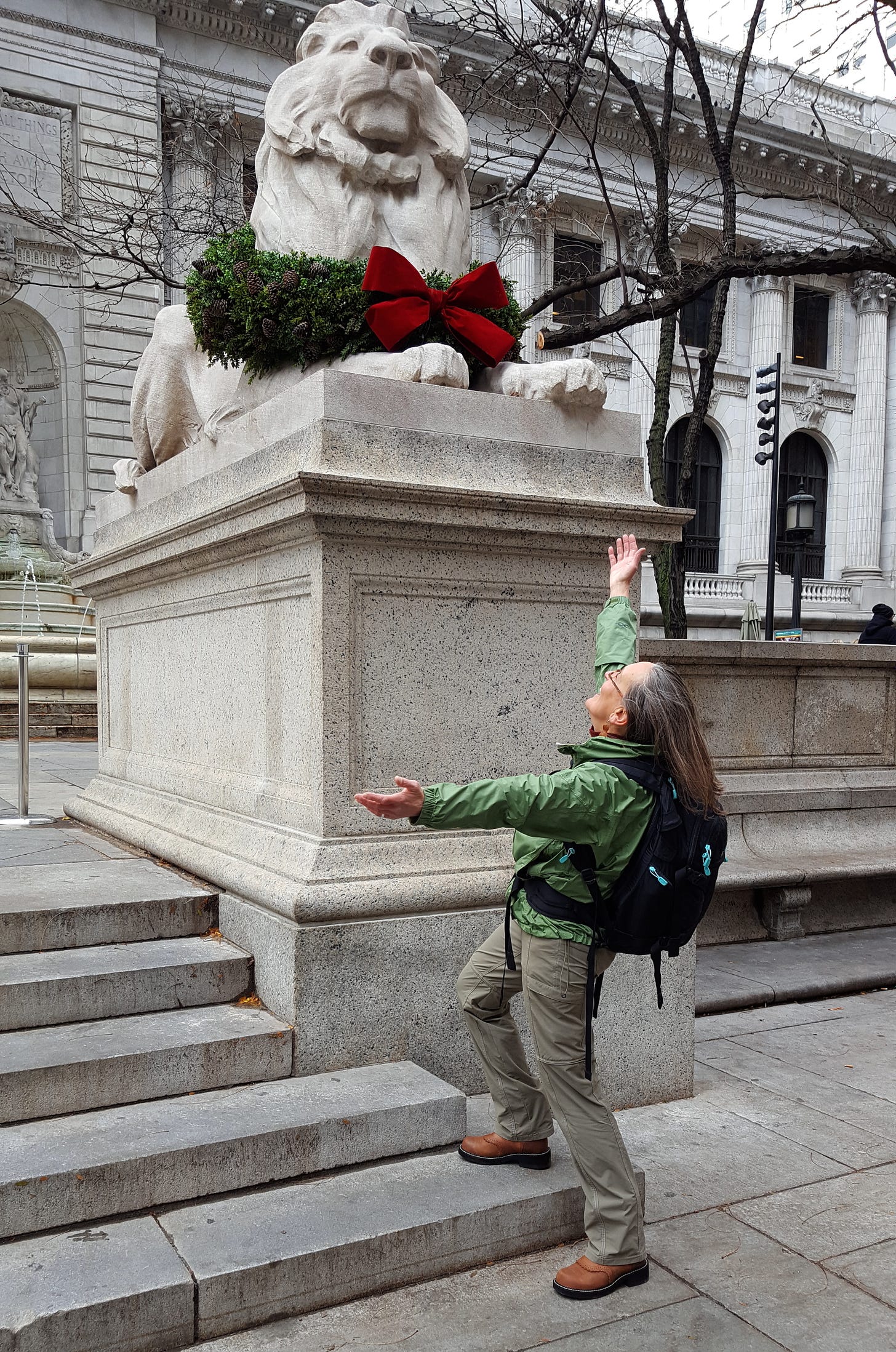 Jodi with long brown hair wearing cowboy boots, a green jacket and a backpack stands at the base of an iconic New York Public Library lion statue — her arms outstretched in homage.