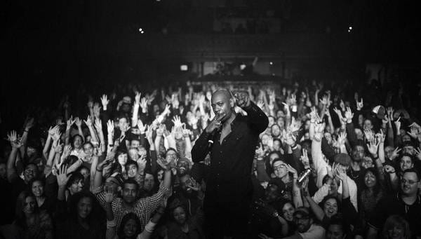 Dave Chappelle stands in front of a large cheering crowd.