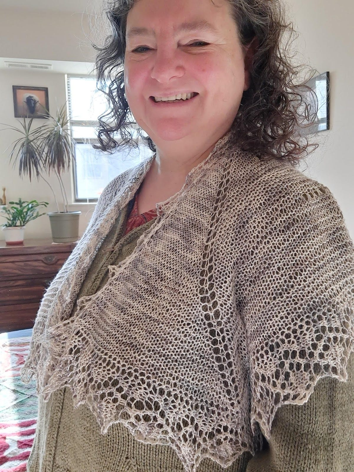 Brown curl haired woman is wearing a gray and white shawl. In the background is a picture of a sheep and some plants.