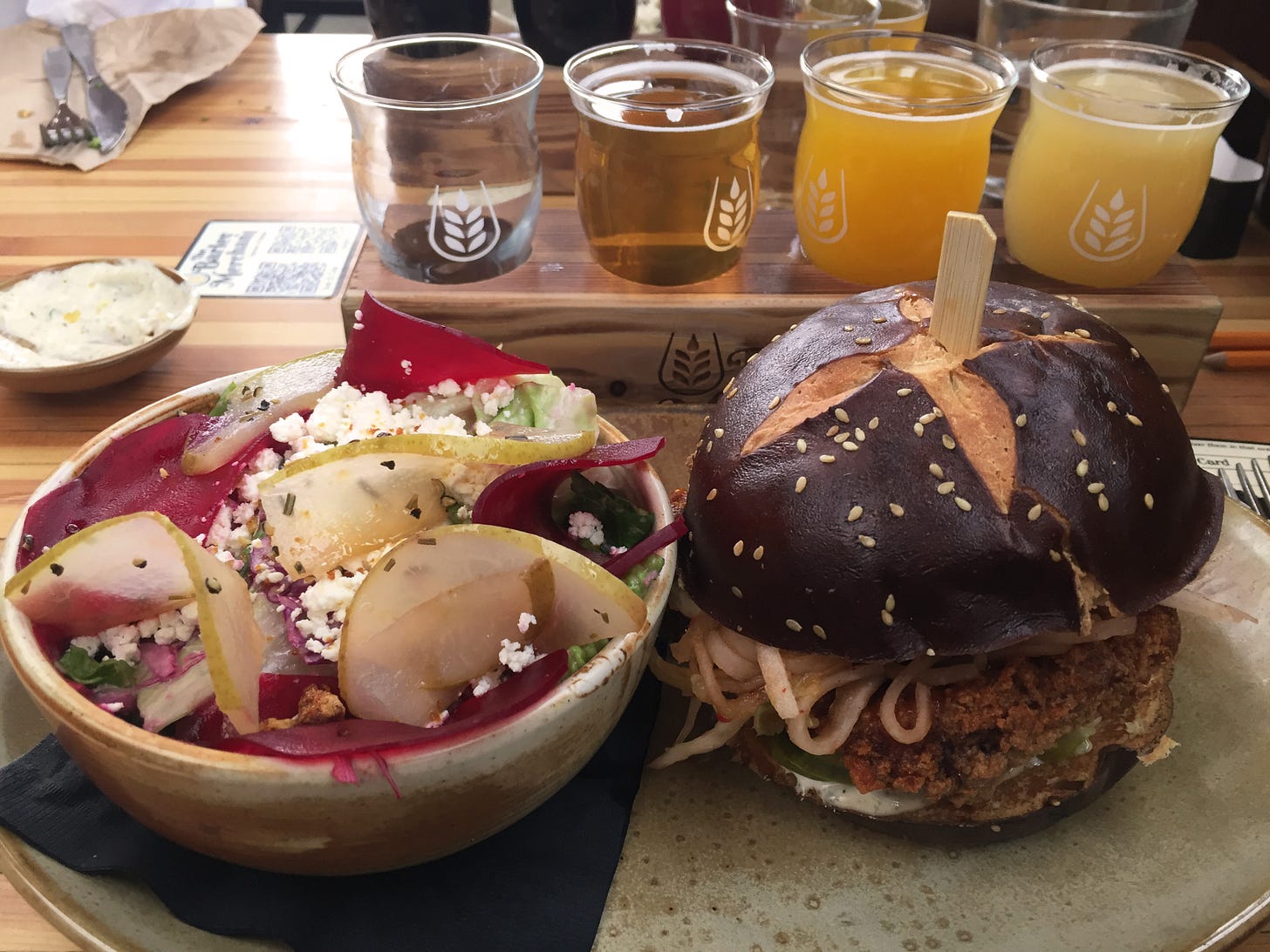 On a wide stone plate, a small bowl of salad with slices of beet and pear and crumbles of goat cheese, beside a fried chicken sandwich with kimchi and pickles on a pretzel bun. Behind the plate is a flight of four beers, one of which is already empty.